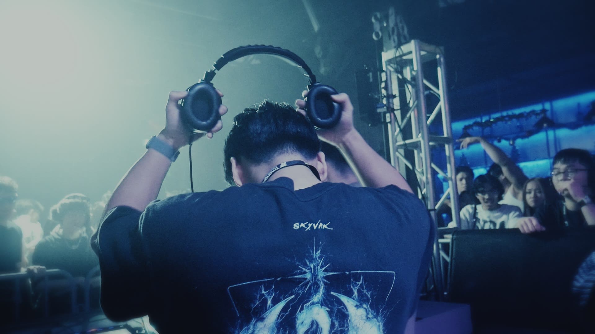 Steven pulling his headphones on behind the booth, crowd out of focus behind him